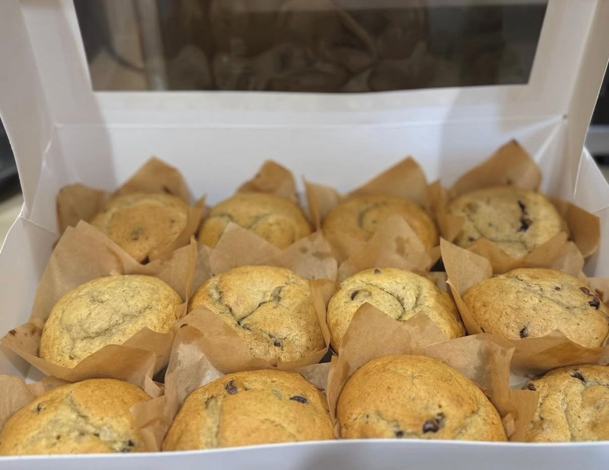 A white bakery box filled with golden-brown chocolate chip muffins in brown paper cups, photographed from above.